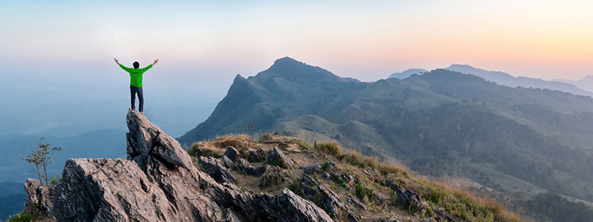 Man in green shirt on high mountain peak at sunrise with his arms up in the air